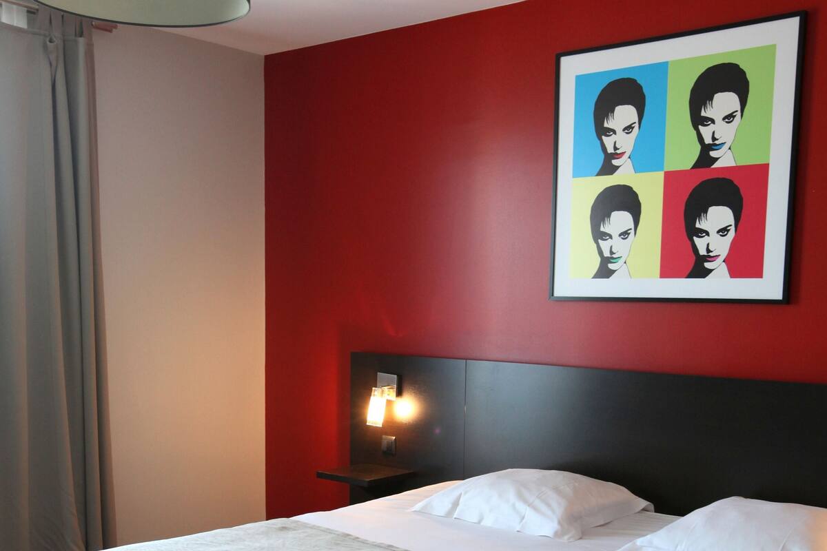 A hotel room with a large bed, a red accent wall, a black headboard, and a modern pendant light.