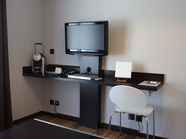 Habitación de hotel moderna con escritorio, televisor de pantalla plana, cafetera, teléfono y una pequeña silla blanca.