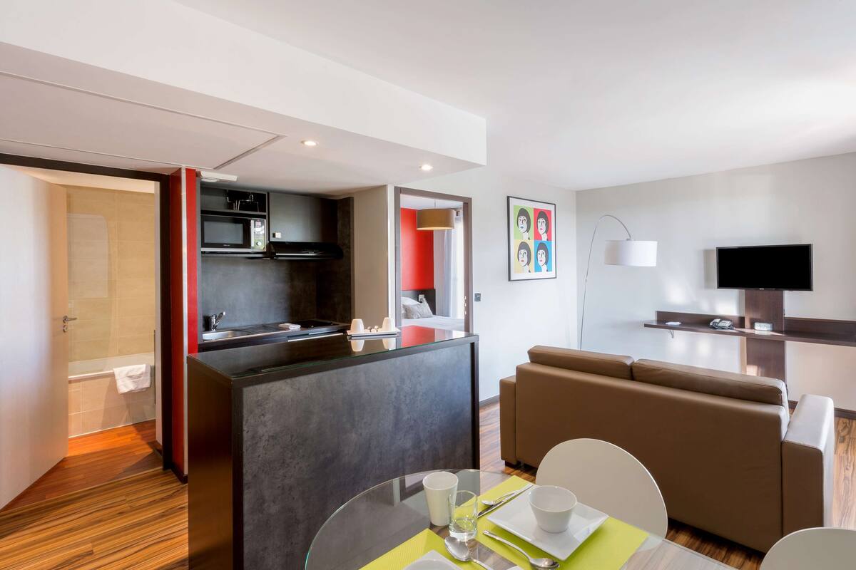 A modern hotel room with a kitchenette, a dining area, a sofa, and a TV.