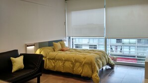 A bedroom with a bed, a black leather sofa, and a window with blinds.