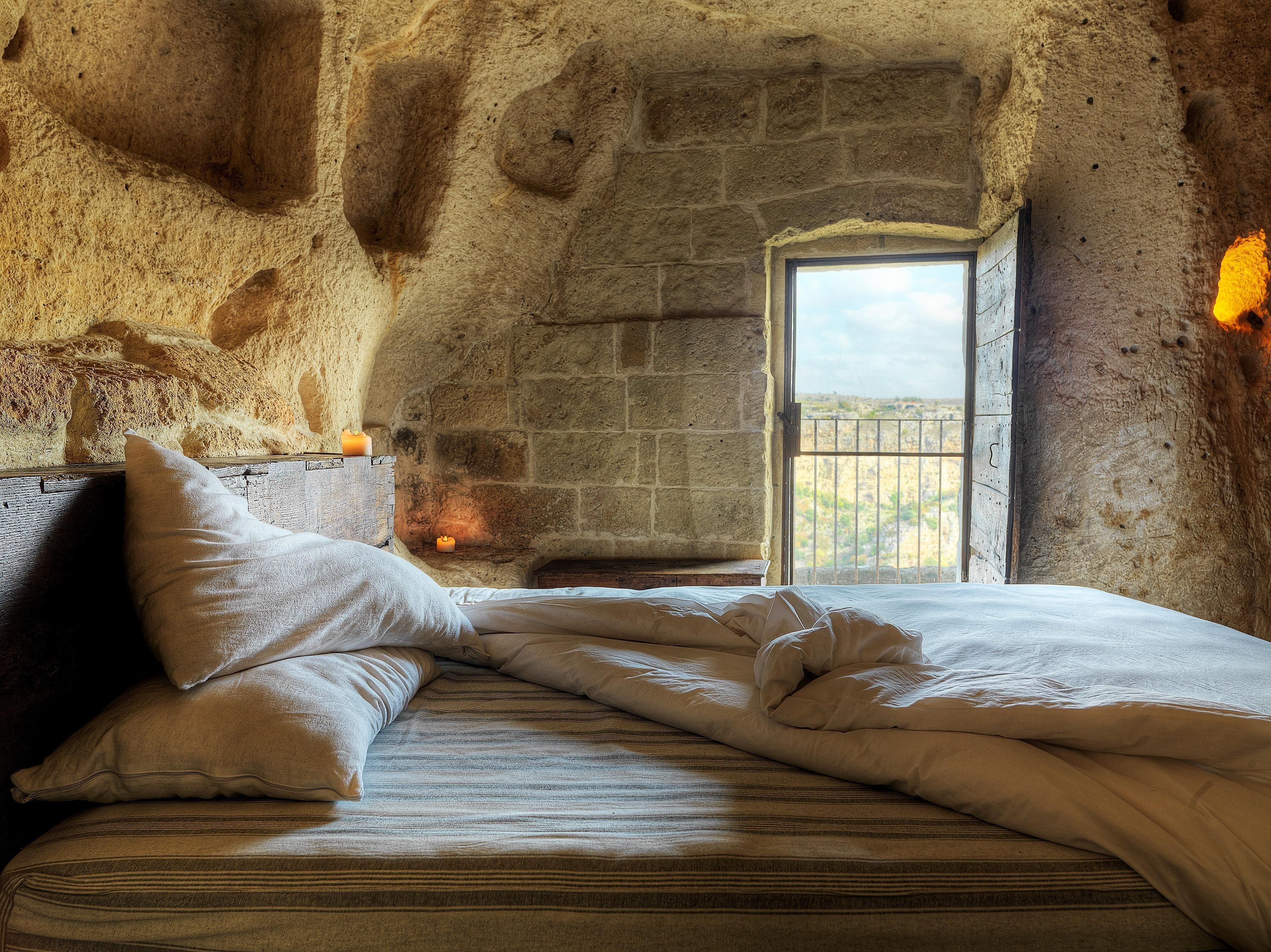 Habitación rústica con paredes de piedra, una cama, almohadas y una ventana con vistas al paisaje.