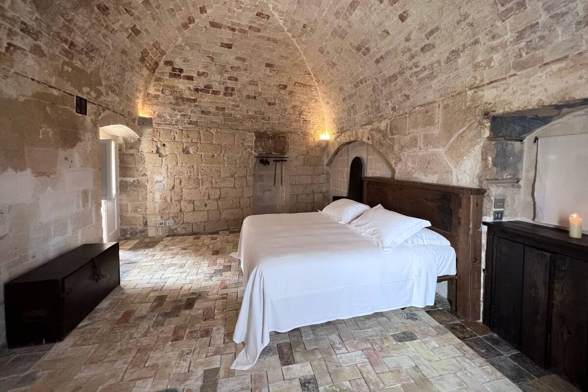 Une pièce avec un plafond et des murs en pierre, un lit avec du linge de maison blanc, une tête de lit en bois, une petite table avec une bougie et un sol en pierre.