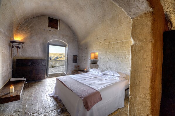 Un dormitorio rústico con paredes de piedra, una cama, una chimenea y vistas a través de un arco abierto.