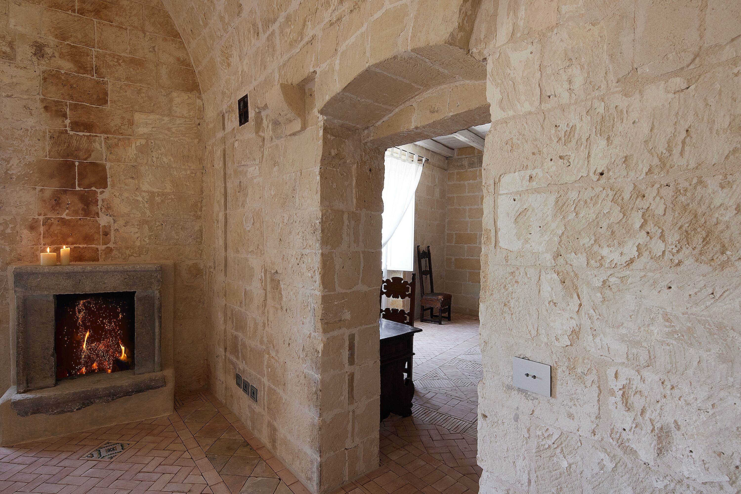A stone-walled room with a fireplace, a wooden chair, and a patterned floor.