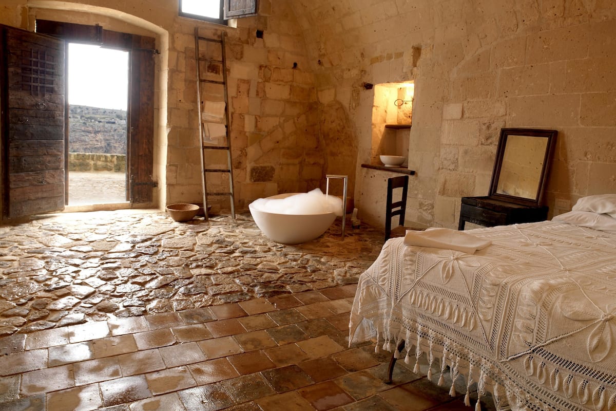 Une chambre rustique avec un sol en pierre, une grande baignoire blanche, un lit en bois et une petite table avec un miroir.