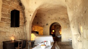Una habitación con paredes de piedra, una cama, una mesa con un frutero y una silla de madera.