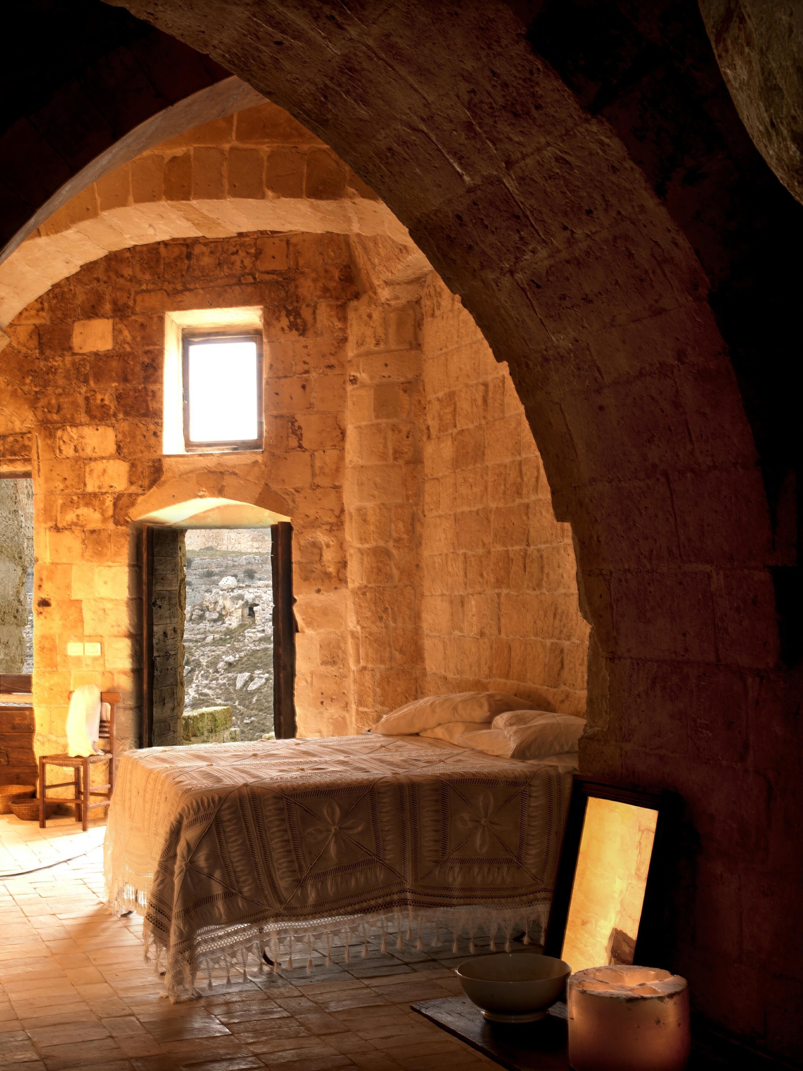 A rustic stone room with a bed, a small window, and a stone wall.