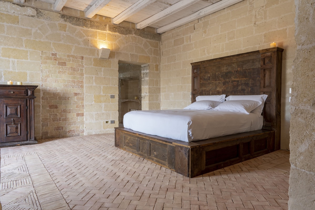 Une chambre avec un lit en bois, des murs en pierre et un parquet disposé en motif à chevrons.