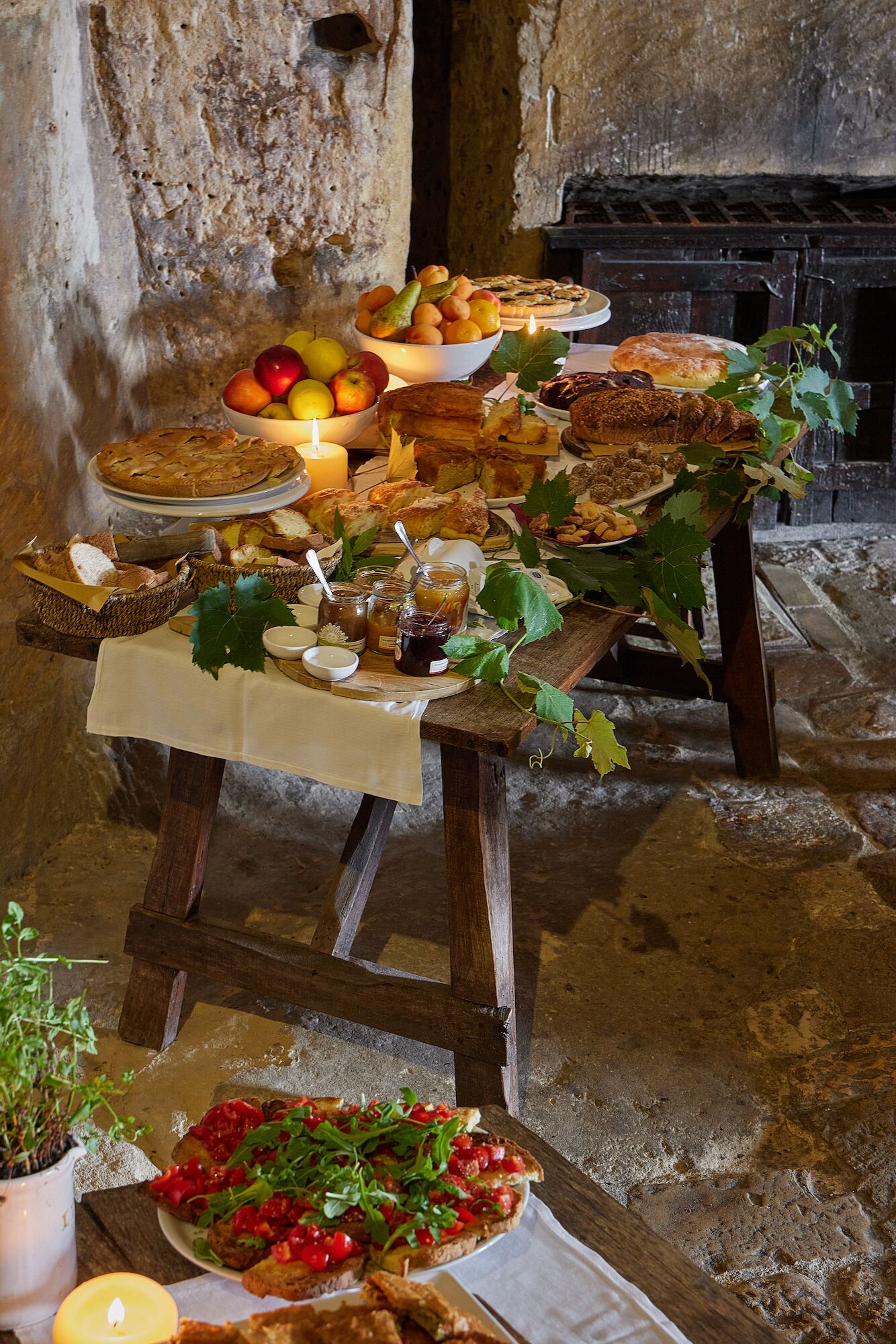 Una mesa de madera rústica con diversos panes, pasteles y frutas, frente a un muro de piedra.
