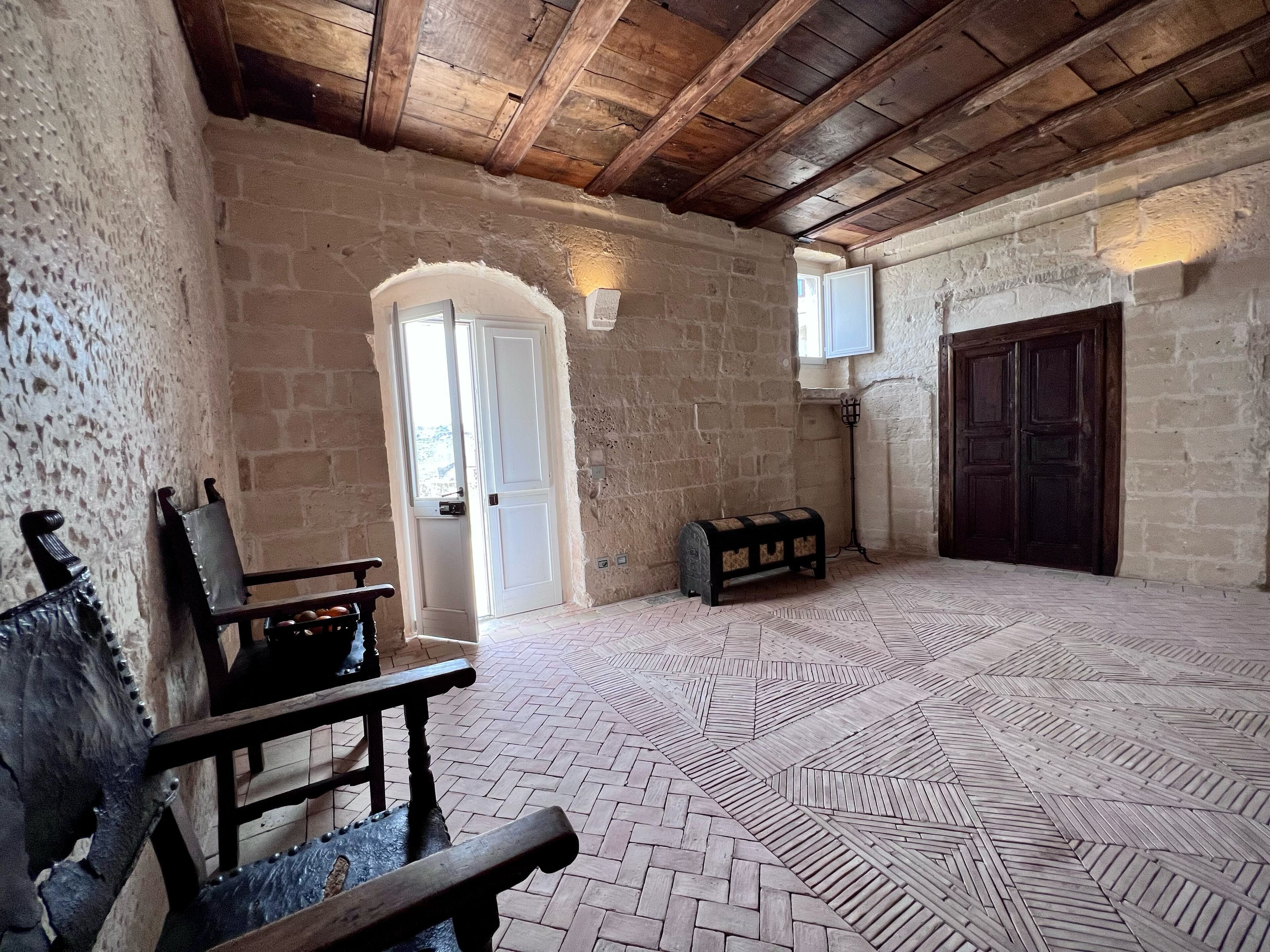 Una habitación con pared de piedra, vigas de madera, un banco, una puerta y suelo estampado.