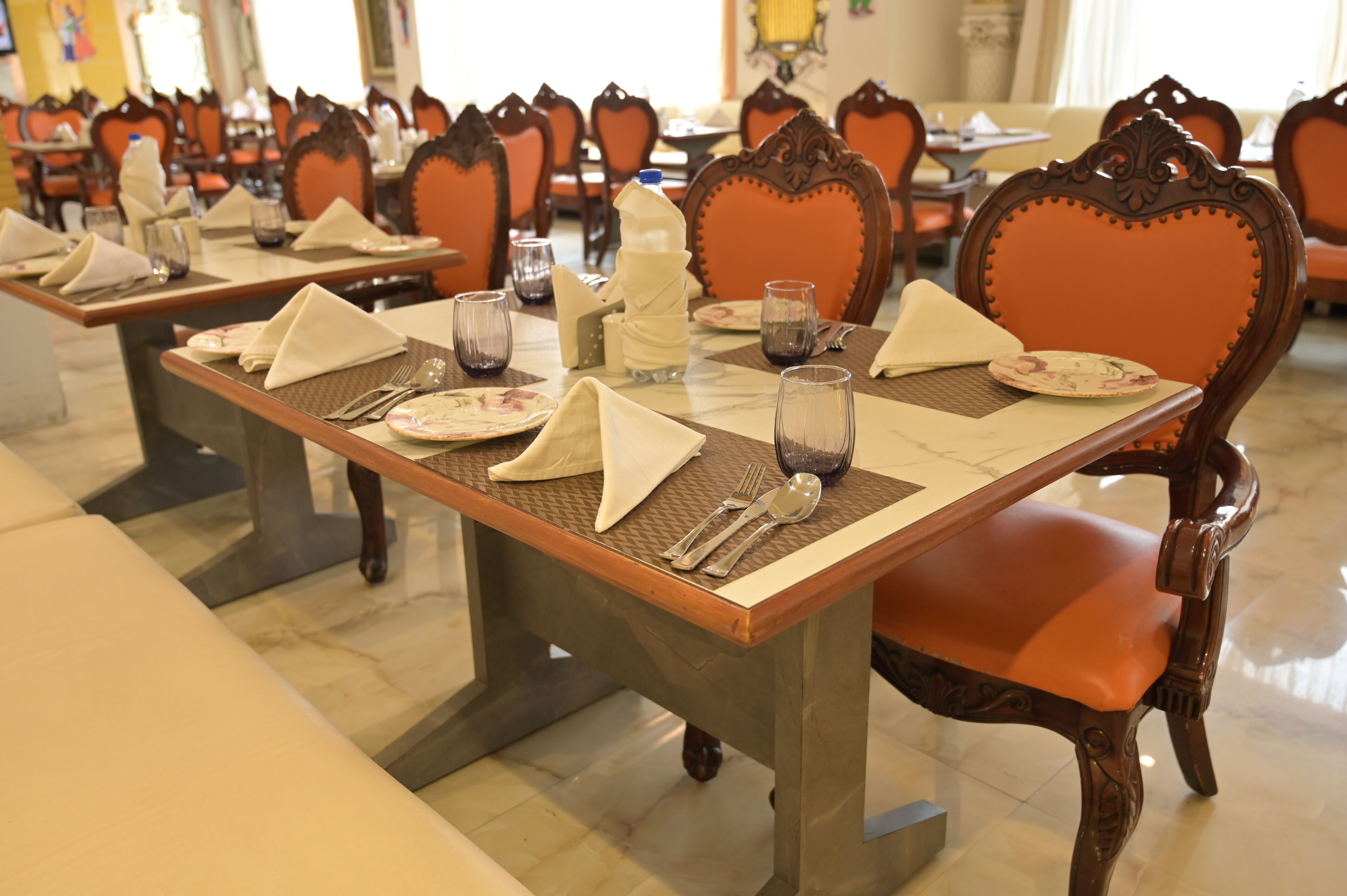 Un comedor con mesas dispuestas para el servicio, sillas con respaldos decorados y un suelo pulido.
