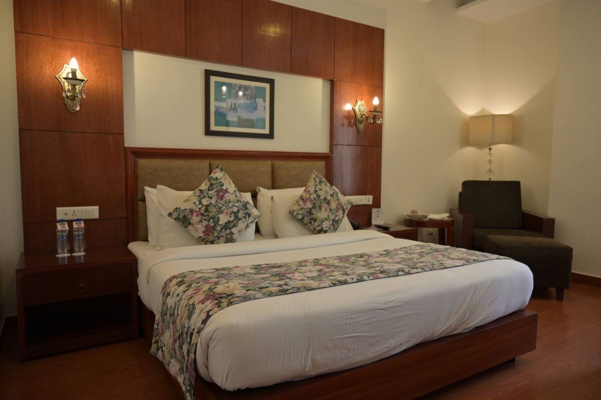 A hotel room with a large bed, wooden headboard, floral pillows, a framed picture above the bed, bedside lamps, a chair, and a nightstand with a bottle and glasses.
