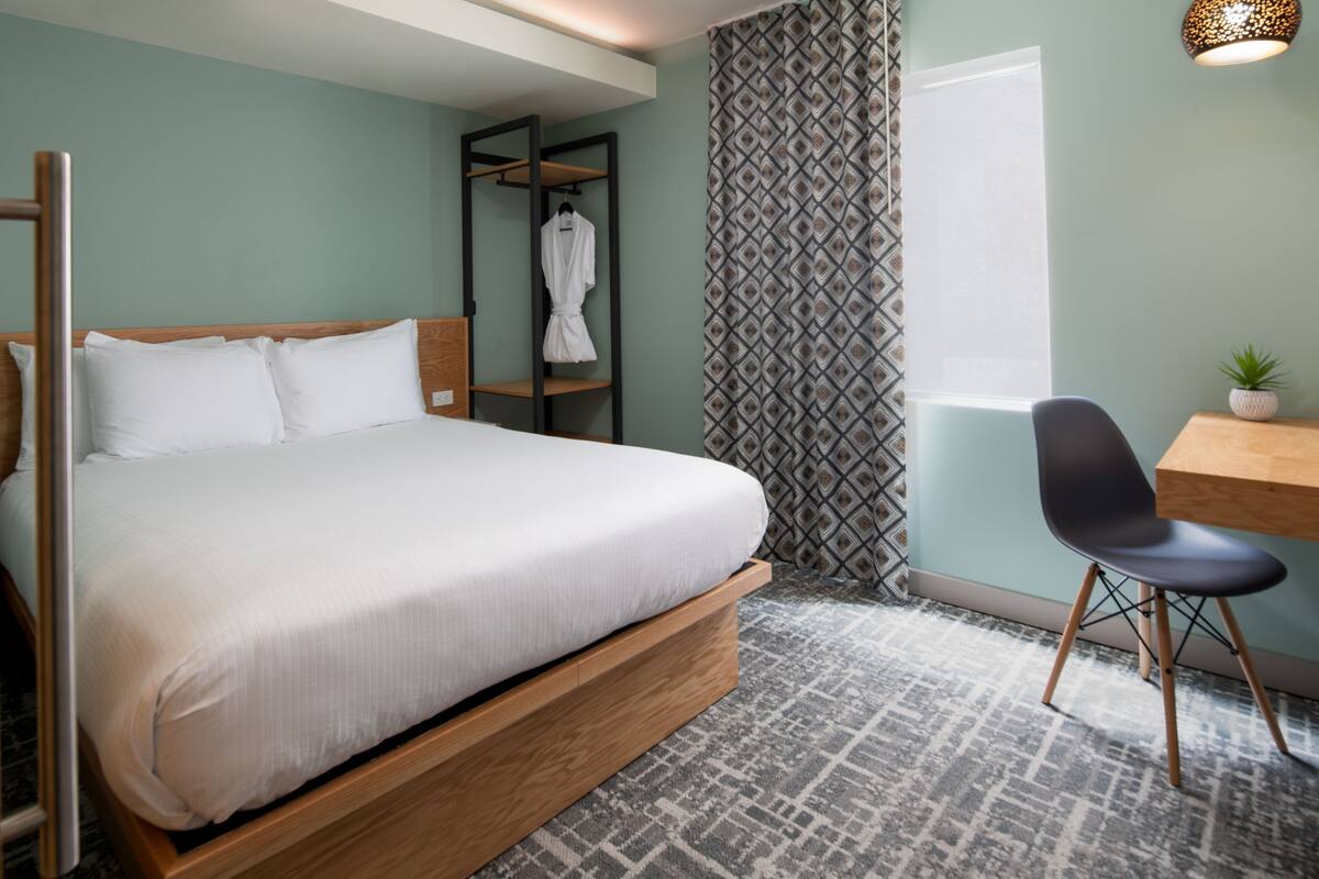 A modern bedroom with a wooden bed, a small desk, a chair, a robe hanging on a rack, and a patterned curtain.