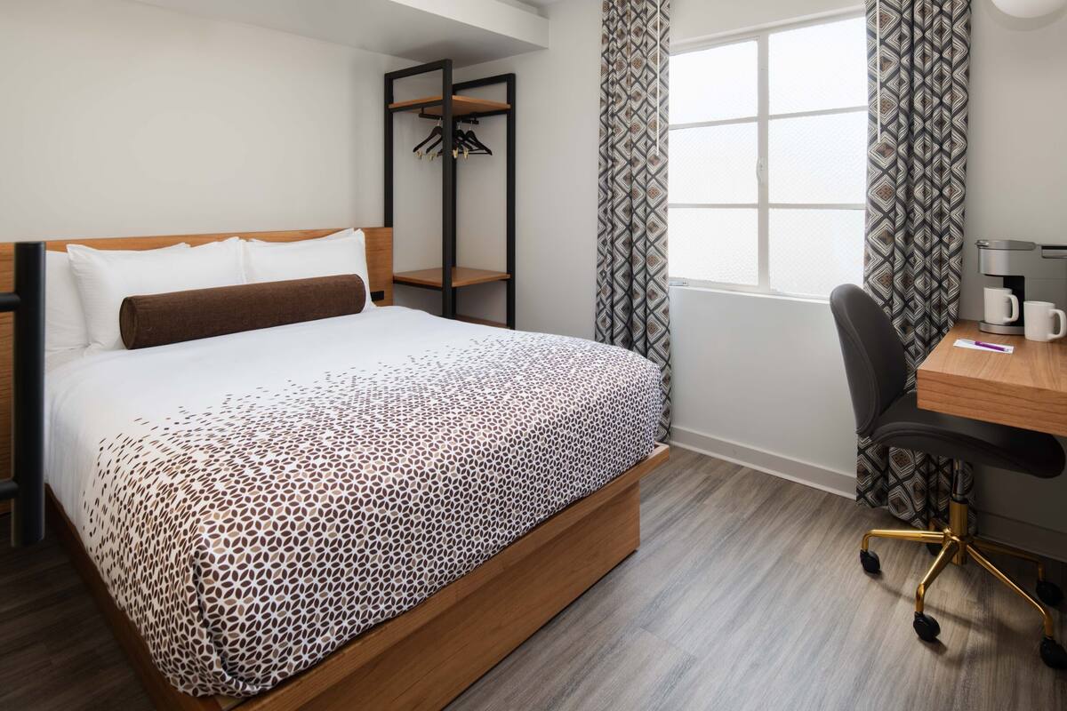 A hotel room with a bed, a desk, a chair, a coffee maker, and a window with curtains.