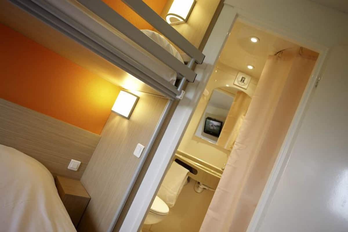 Una habitación de hotel compacta con una cama litera, una mesita de noche pequeña, un televisor montado en la pared y un baño con inodoro y lavabo.