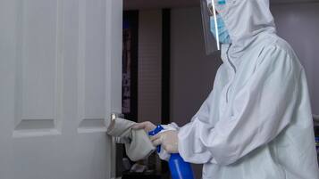 Una persona con traje y careta de protección blancos, sosteniendo una botella con atomizador azul, está de pie en un umbral.