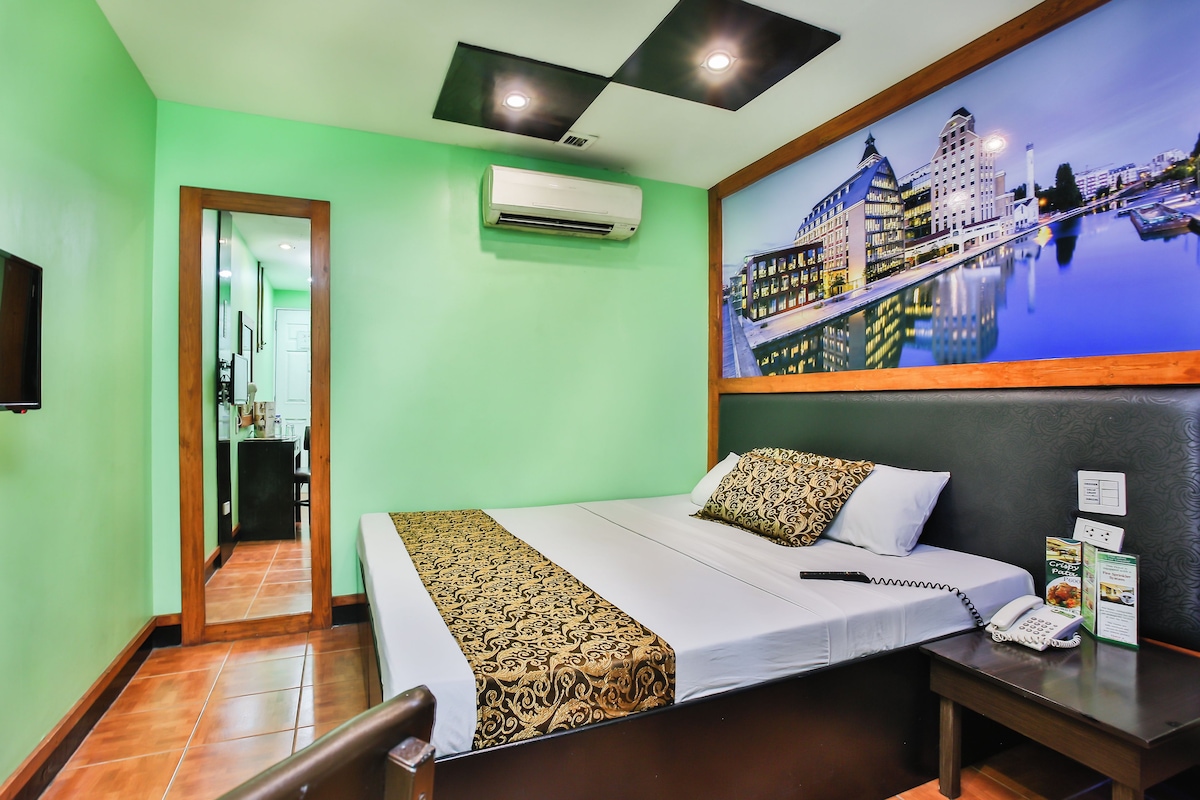Habitación de hotel con cama, mesita de noche, aire acondicionado instalado en la pared, un cuadro enmarcado con una vista urbana y una puerta de madera que da a un pasillo.