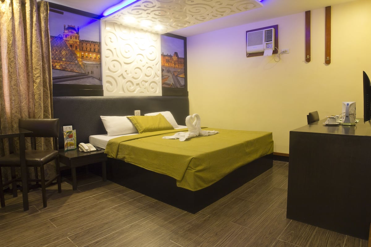 A hotel room with a bed, a desk, a chair, and a wall-mounted air conditioner.