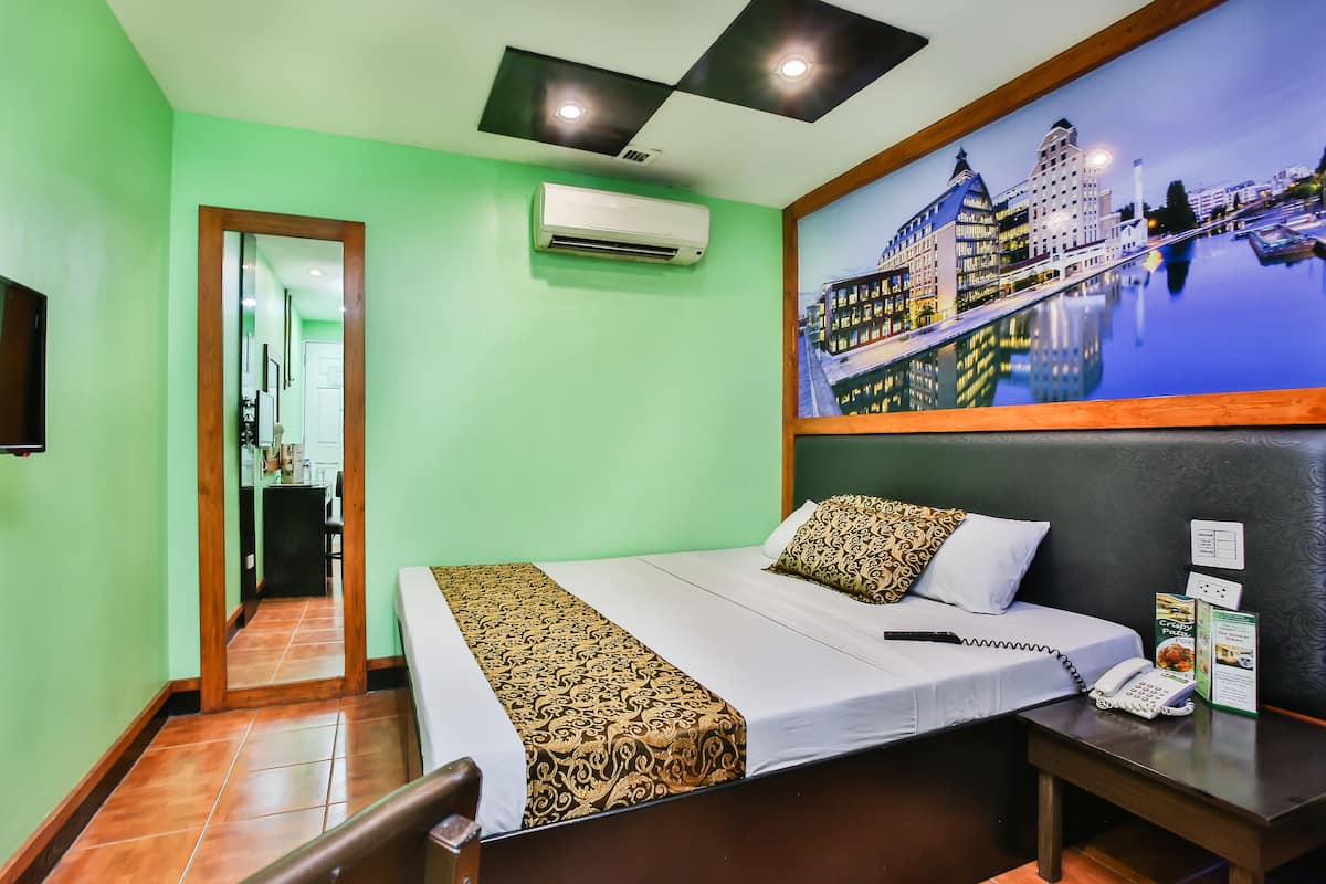 A hotel room with a bed, a desk with a telephone, an air conditioning unit, and a painting of a cityscape.