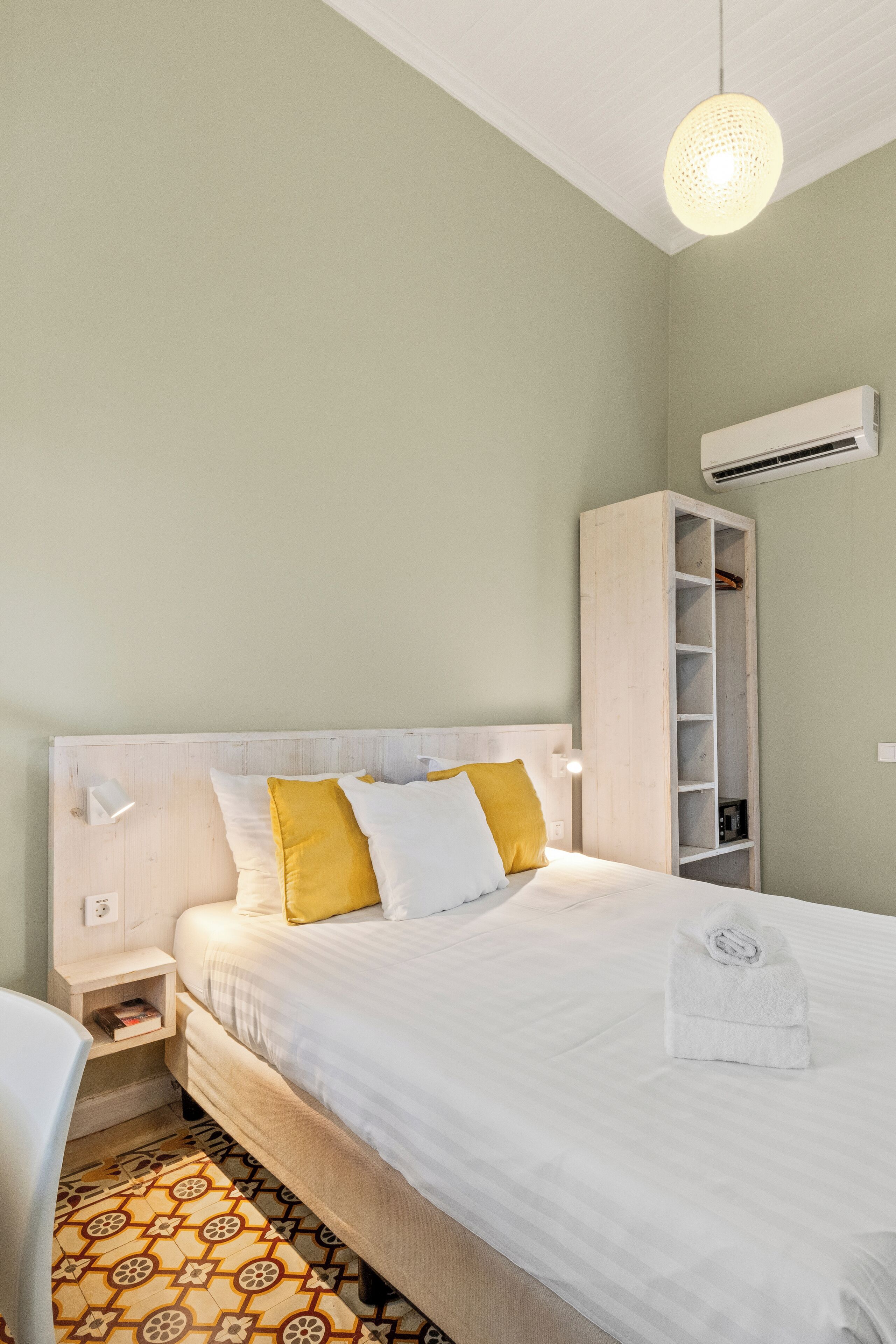 Een net opgemaakt bed met witte en gele kussens, een nachtkastje met een lamp en een inbouwkast met planken.