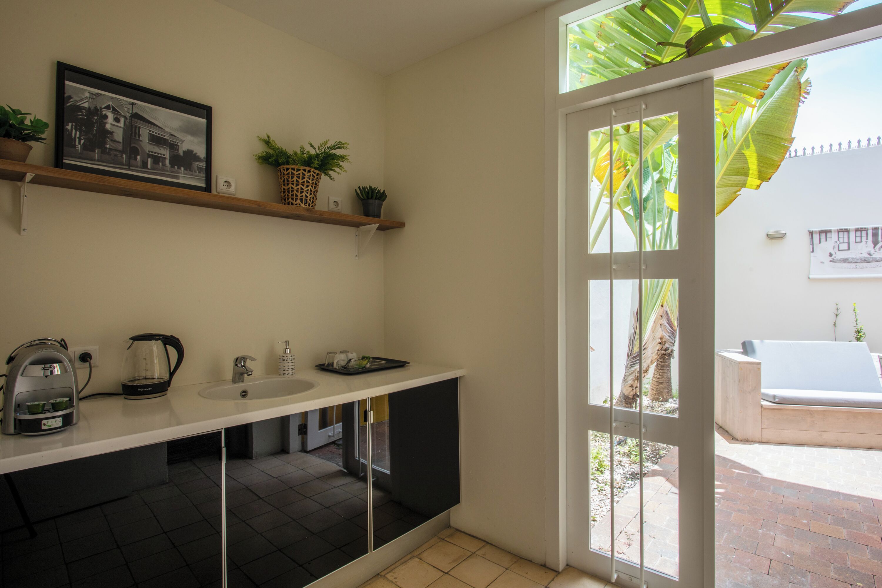 A modern kitchen with a white countertop, black cabinets, a coffee maker, and a sink. There is a window with a view of a patio and a white bathtub.