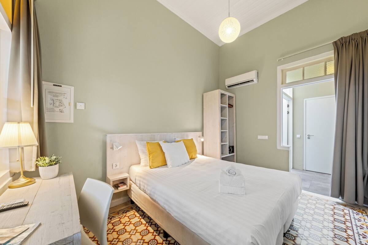 A hotel room with a bed, a desk with a lamp, a plant, and a patterned floor.