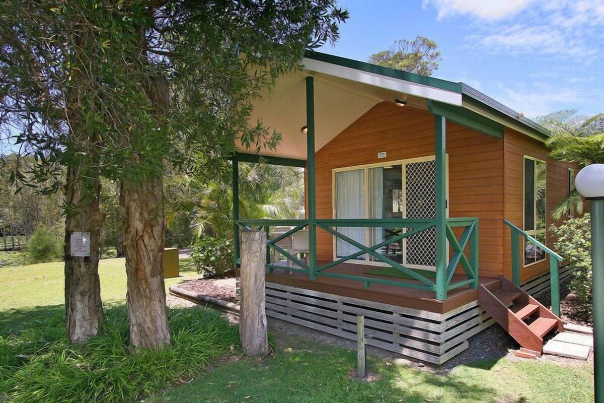 Una pequeña cabaña de madera con un porche, rodeada de árboles y hierba.