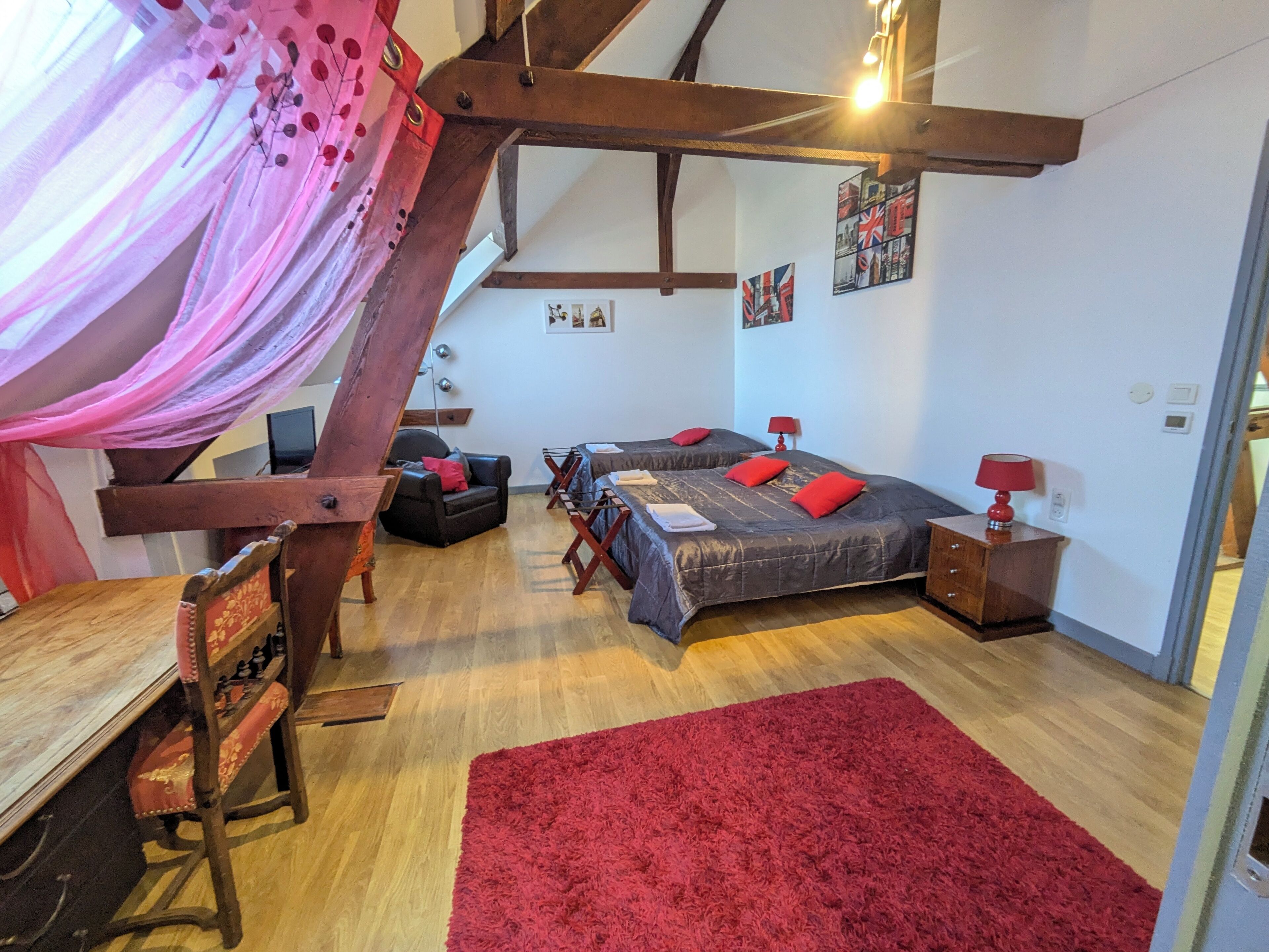 A cozy bedroom with a wooden ceiling, two beds with red pillows, a red rug, and a desk with a chair.