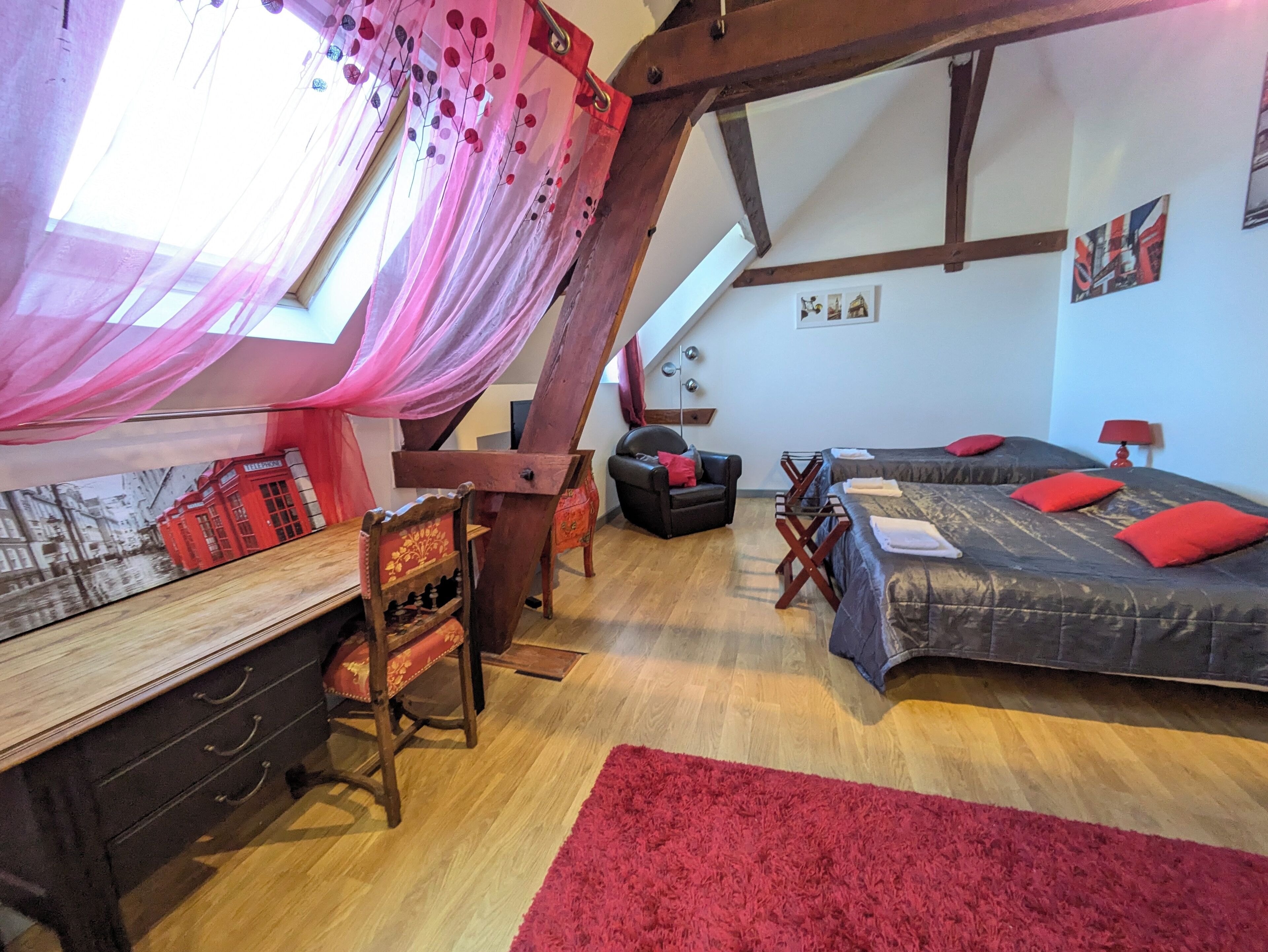 A cozy bedroom with a wooden desk, a bed with red pillows, a red rug, and a sloped ceiling with exposed beams.