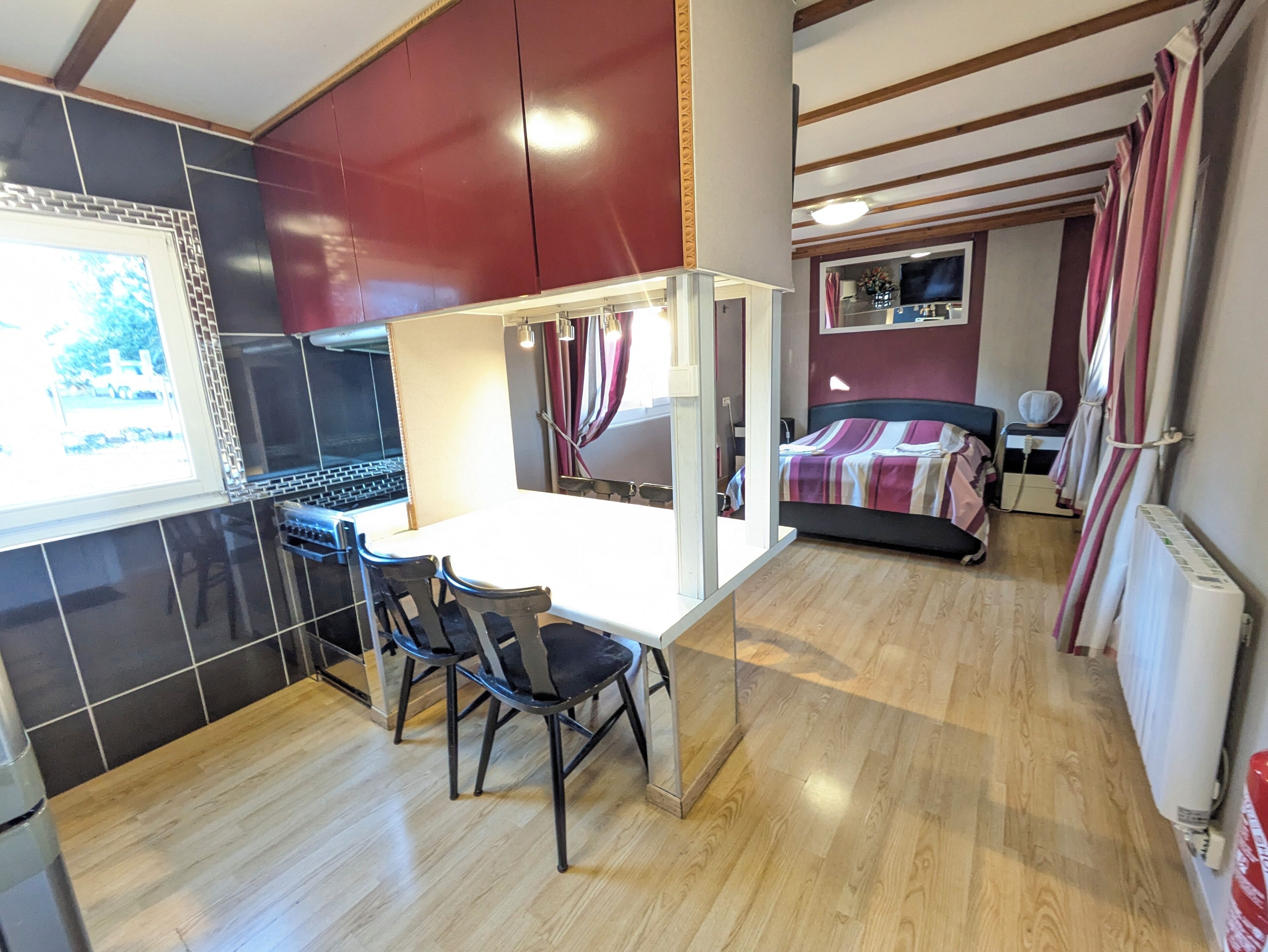 A compact room with a kitchenette, a dining area with two chairs, a bed with a striped blanket, and a large mirror.