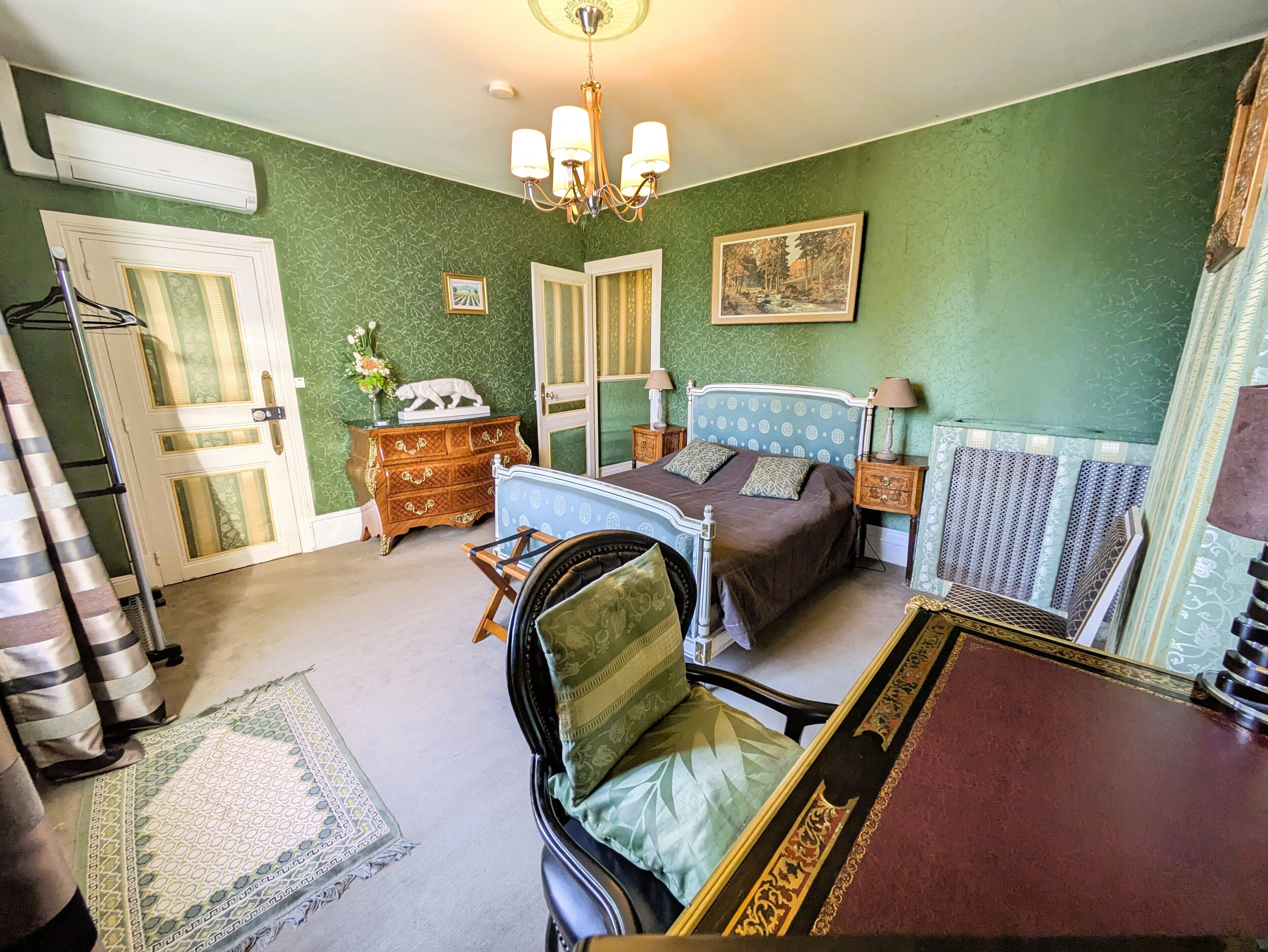 Un dormitorio con cama, cómoda, silla y mesa con otra silla. Se observa una puerta, un radiador y una araña.