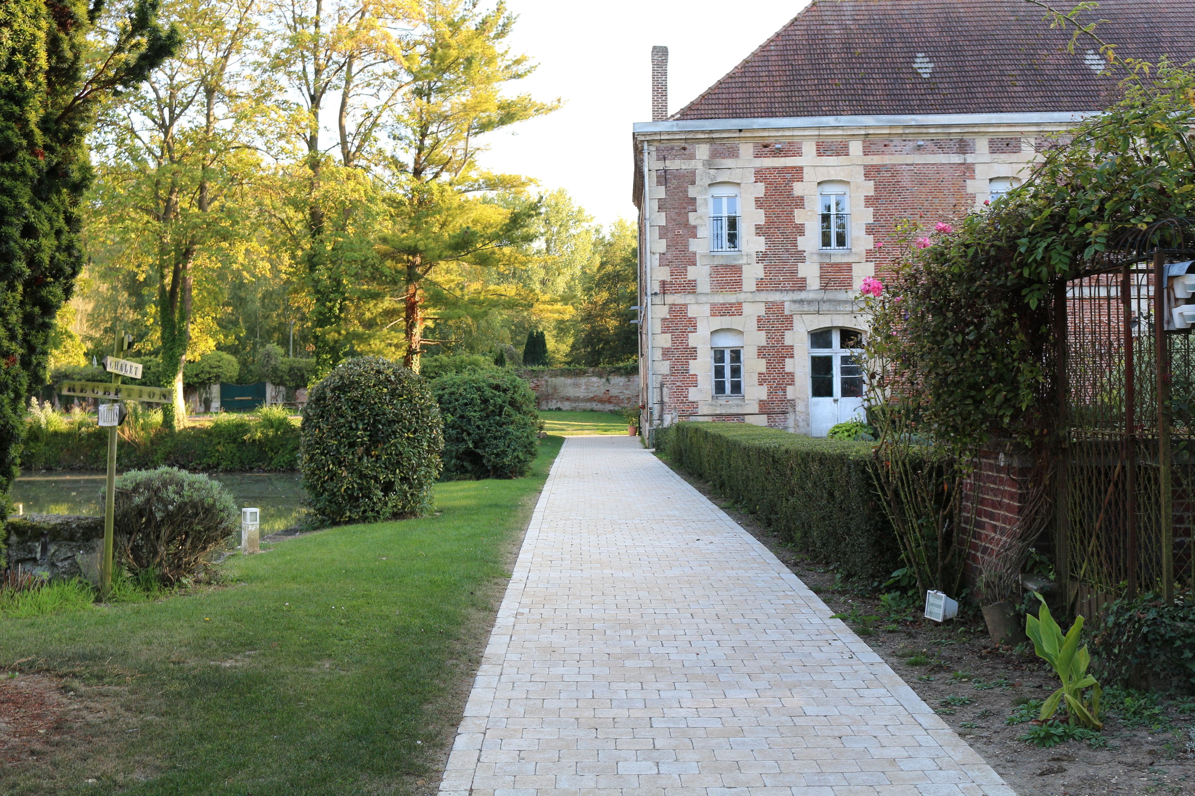 A paved pathway leading to a brick building with a garden and trees.