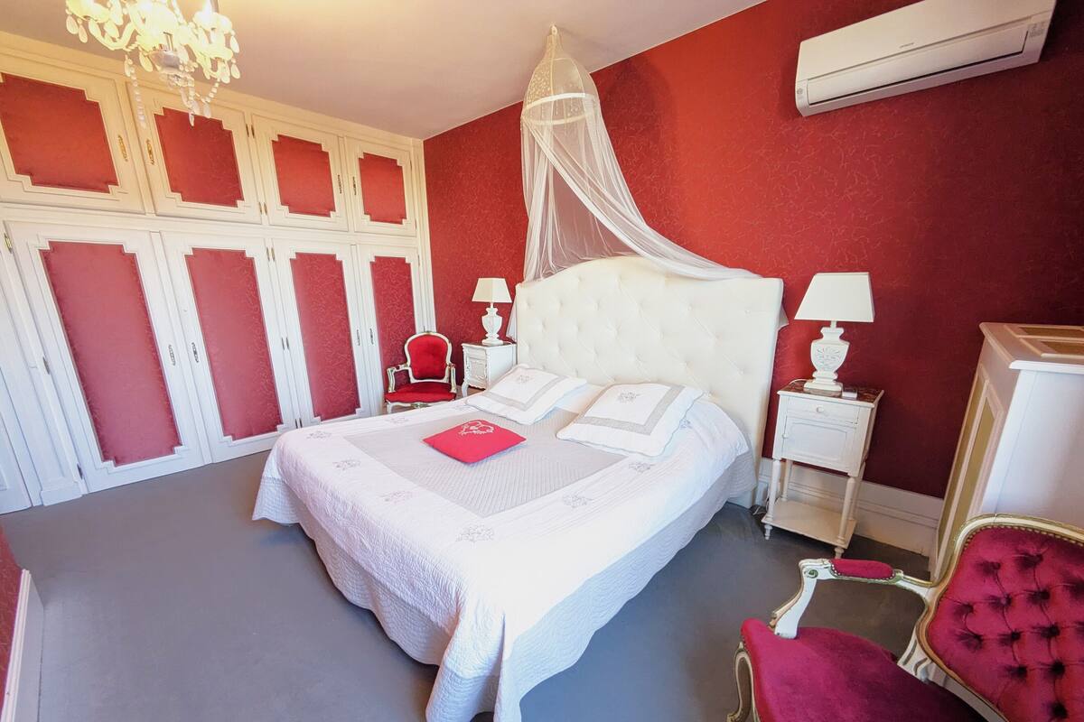 Ein ordentlich eingerichtetes Schlafzimmer mit einem großen Bett, einem roten Sessel, einem weißen Nachttisch und einer Chandelüster.