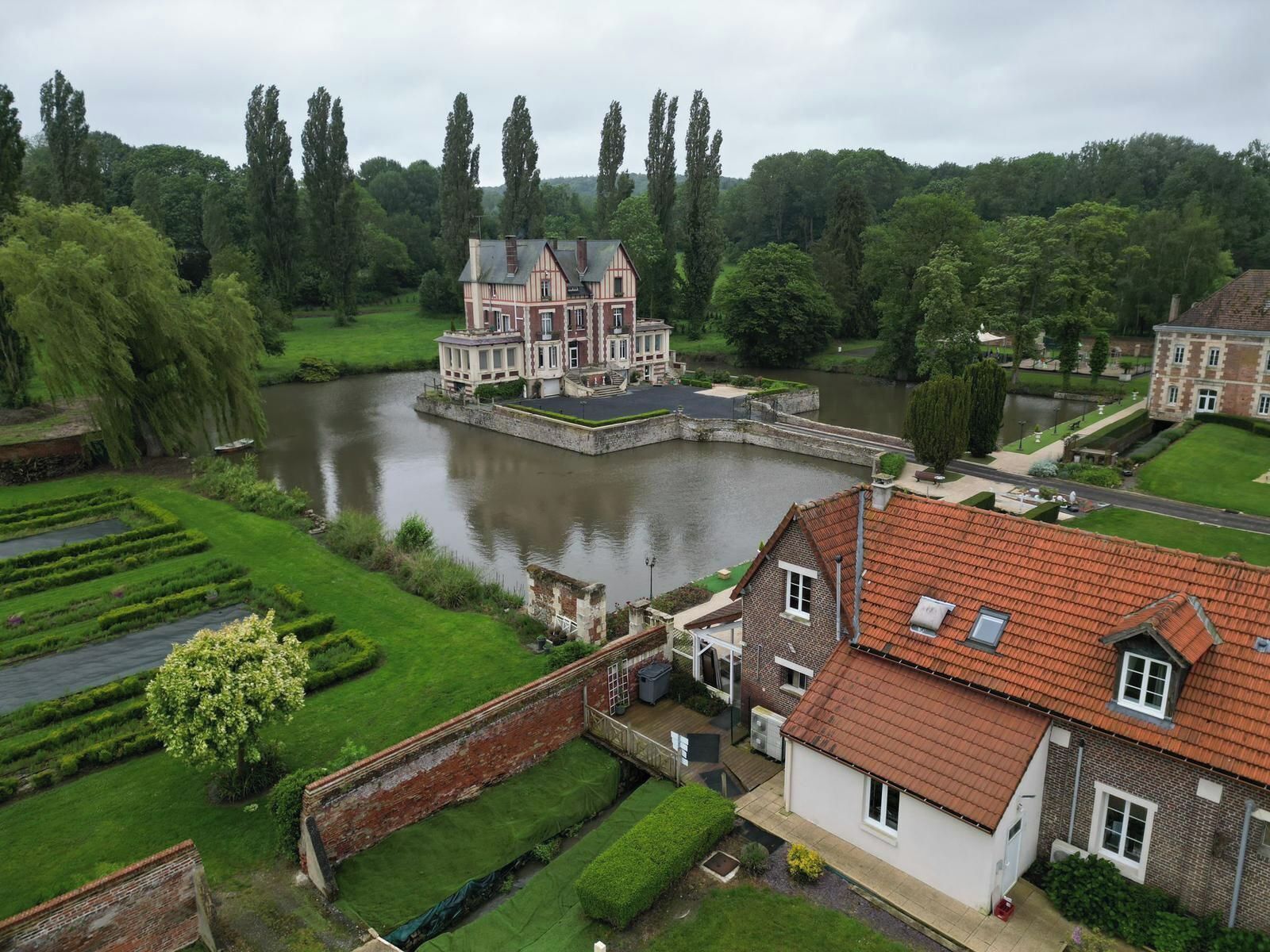 A large, historic building with a river in front, surrounded by lush greenery and tall trees.