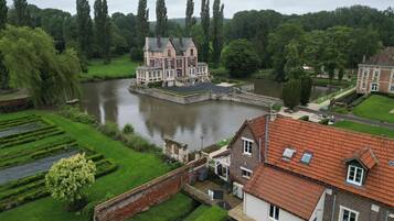 A large, historic building with a river in front, surrounded by lush greenery and tall trees.