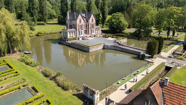 Een groot, historisch gebouw met een symmetrische gevel, gelegen aan een rustig meer, omringd door verzorgde tuinen en paden.