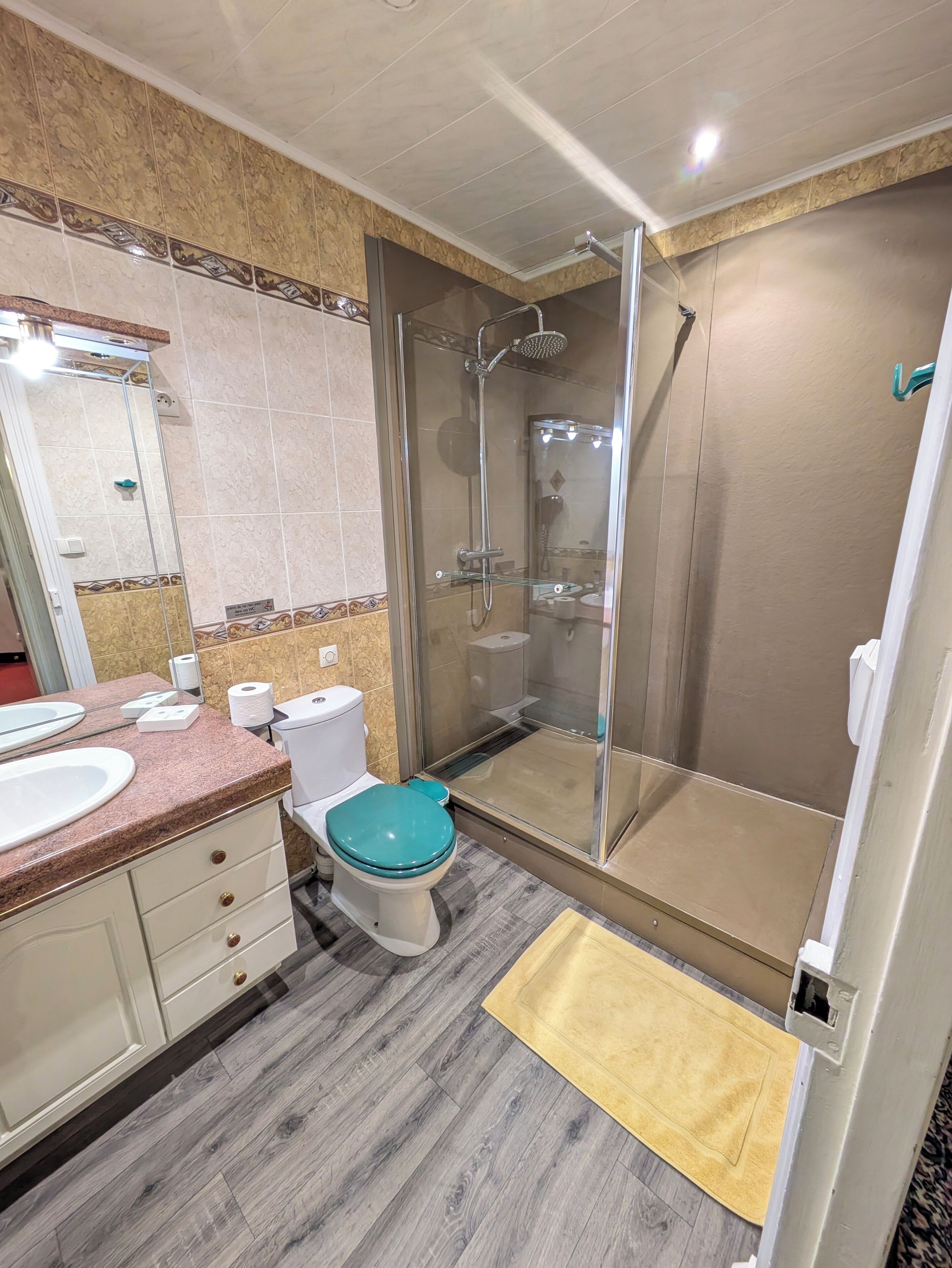 Un bany amb dutxa tancada amb vidre, un vàter amb seient color turquesa, un lavabo i un mirall.