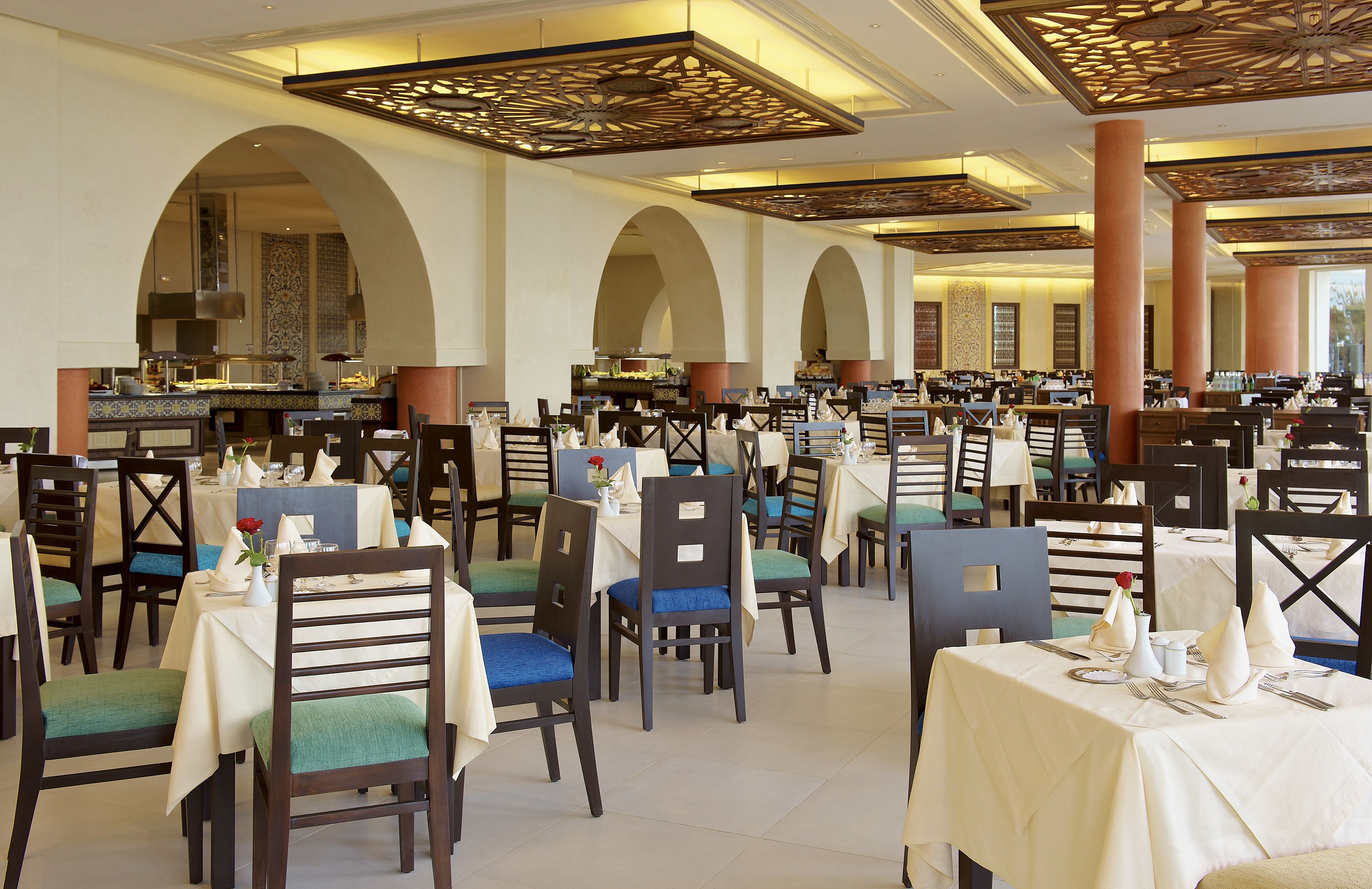 A spacious dining area with multiple tables and chairs, a central bar, and a high ceiling with decorative beams.