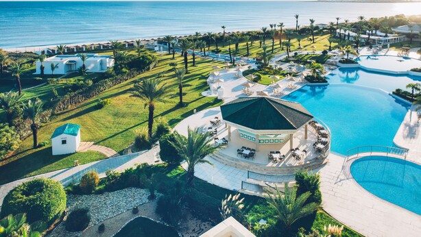 An aerial view of a resort with a large swimming pool, a covered outdoor seating area, and a view of the ocean.