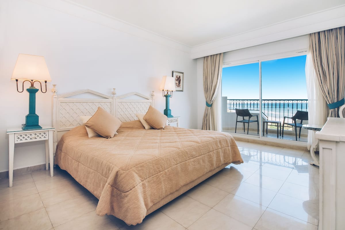 Habitación de hotel con una cama grande, dos mesitas de noche con lámparas, un balcón con sillas y vistas al mar.