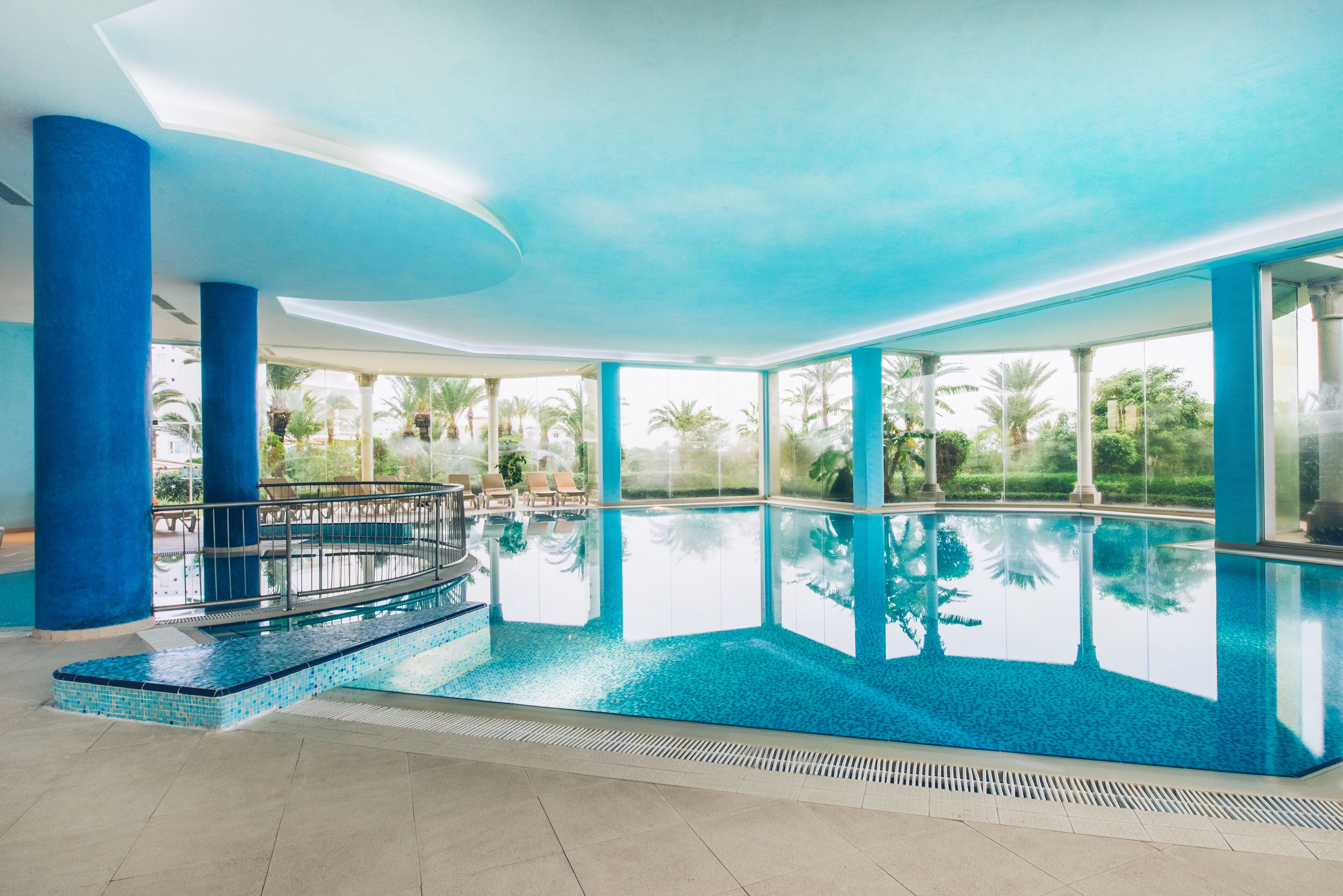 Una piscina interior con agua azul cristalina, rodeada de amplias ventanas y pilares azules.