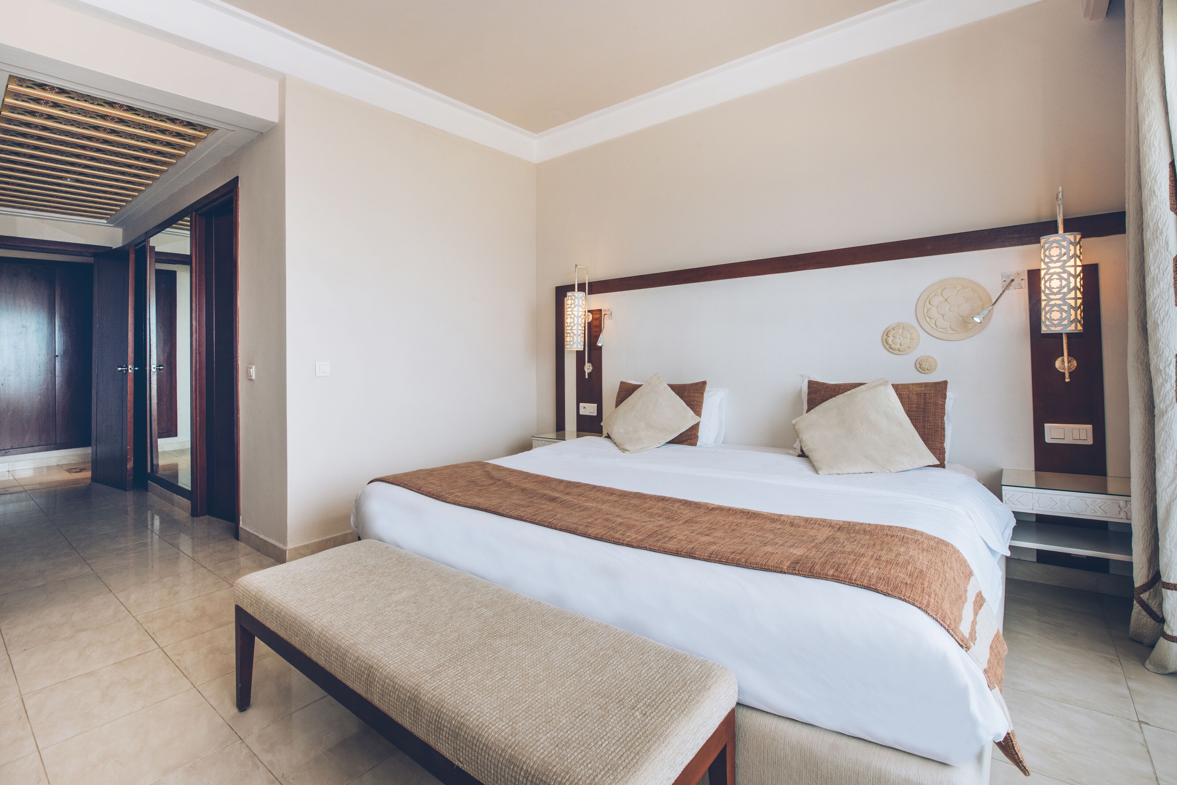 A hotel room with a large bed, a bench, a wooden headboard, and a ceiling light fixture.