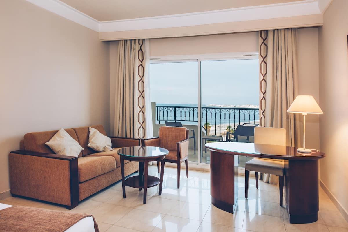 Una habitación de hotel moderna con un sofá, sillas, una mesita y un escritorio con lámpara. La habitación tiene ventanas amplias con cortinas y vista al mar.