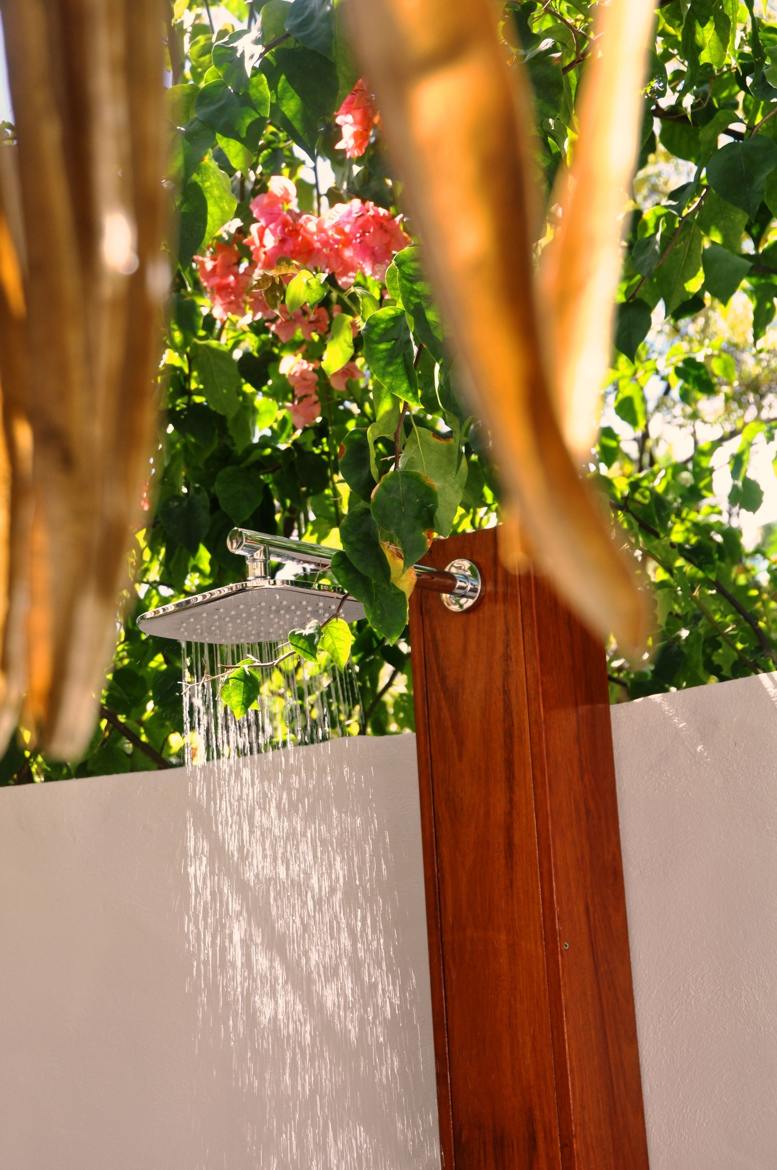 A showerhead with water flowing from it, set against a backdrop of green foliage and pink flowers.