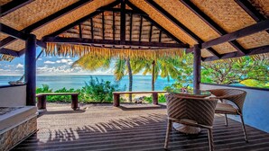 A covered outdoor area with a view of the ocean, featuring a thatched roof, wicker furniture, and a wooden deck.
