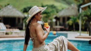 A person sitting by a pool with a drink, wearing a hat and a bikini.