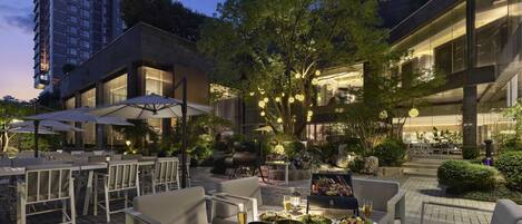 An outdoor dining area with tables and chairs, surrounded by trees and modern buildings.