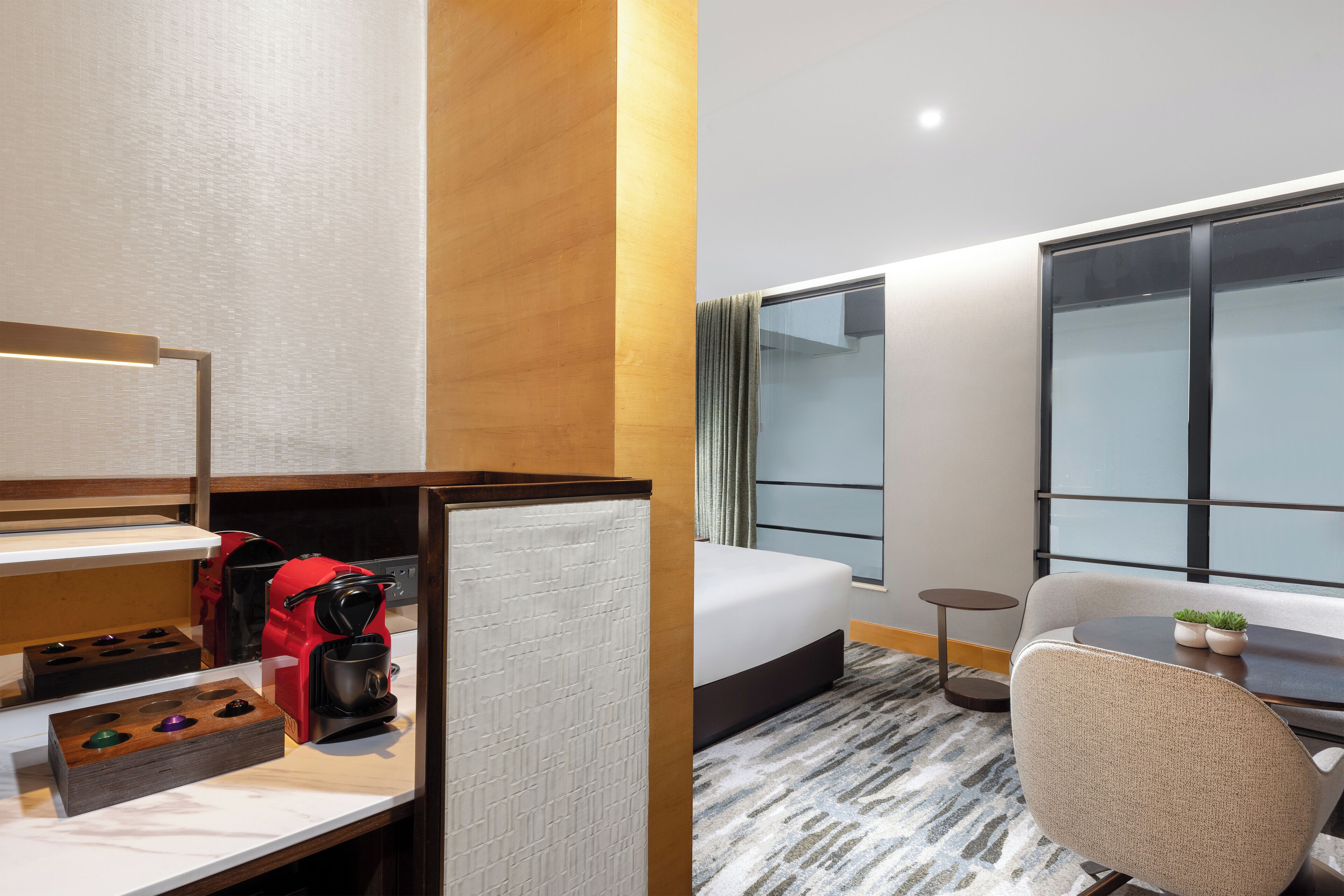 Een hotelkamer met een bed, een bureau met koffiezetapparaat, een stoel, een klein tafeltje met planten en een groot raam.