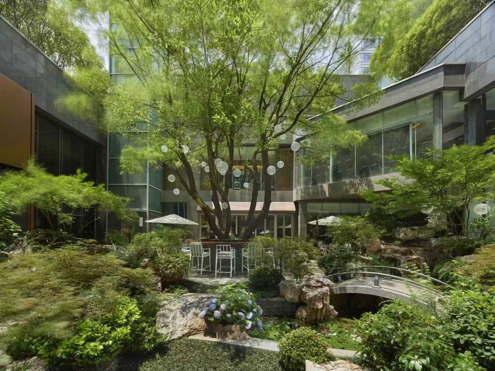 A modern building with a courtyard featuring a tree, a small bridge, and a seating area.
