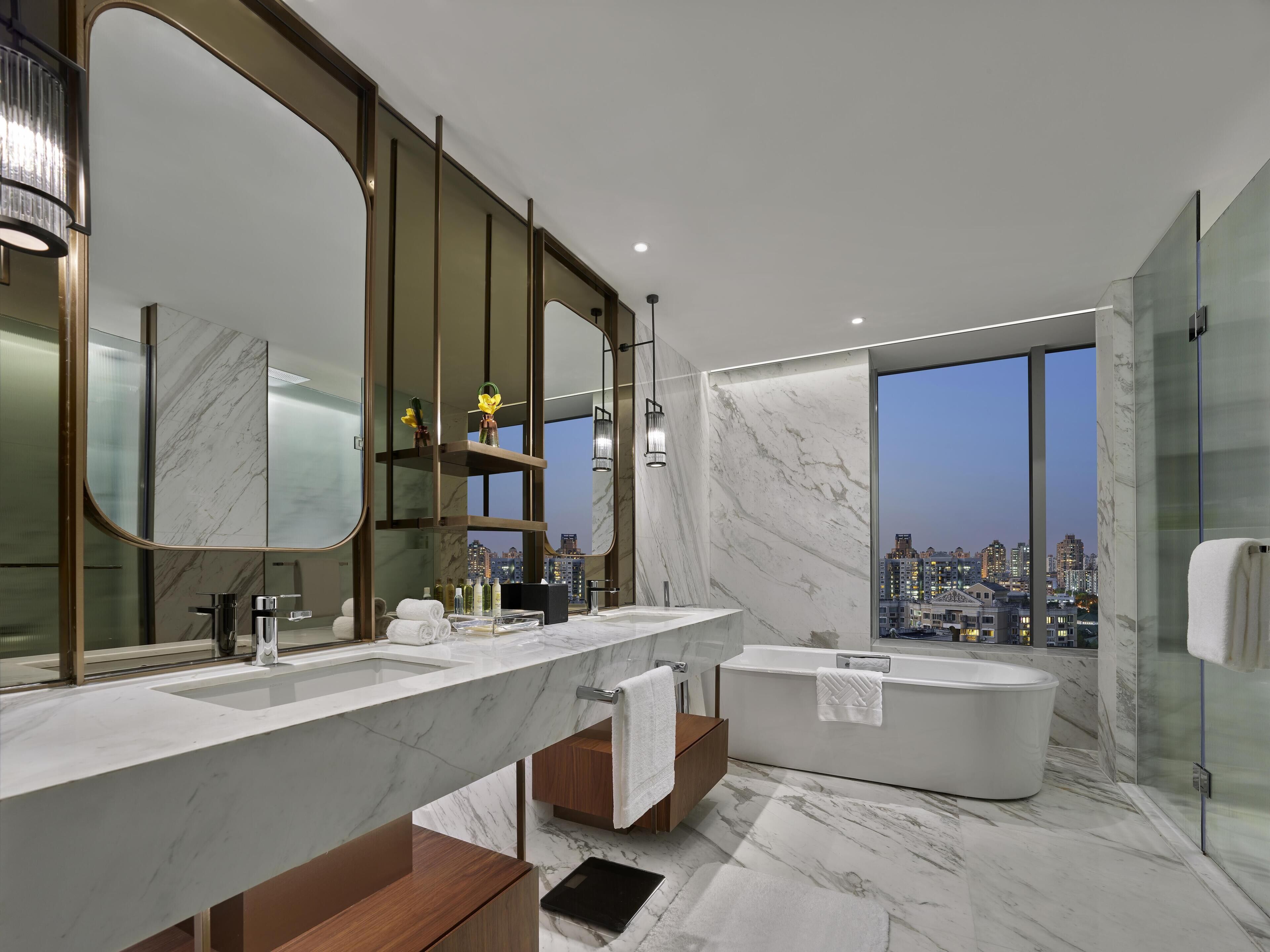 A modern bathroom with a large mirror, two sinks, a bathtub, and a city view.